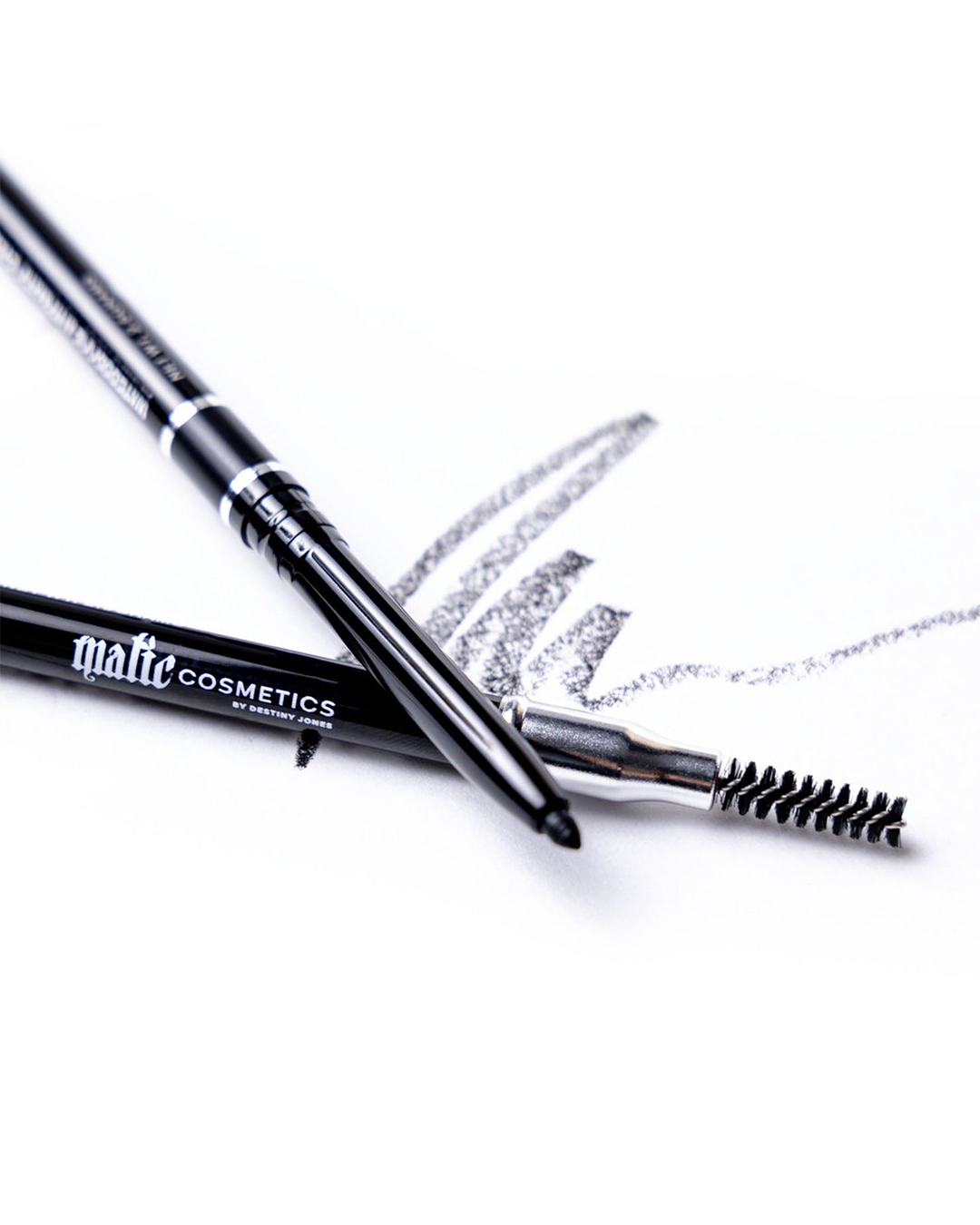 A mechanical eyebrow pencil in soft black color with the brand name visible, alongside an applicator brush, against a white background with a stylized shadow of the pencil and brush.